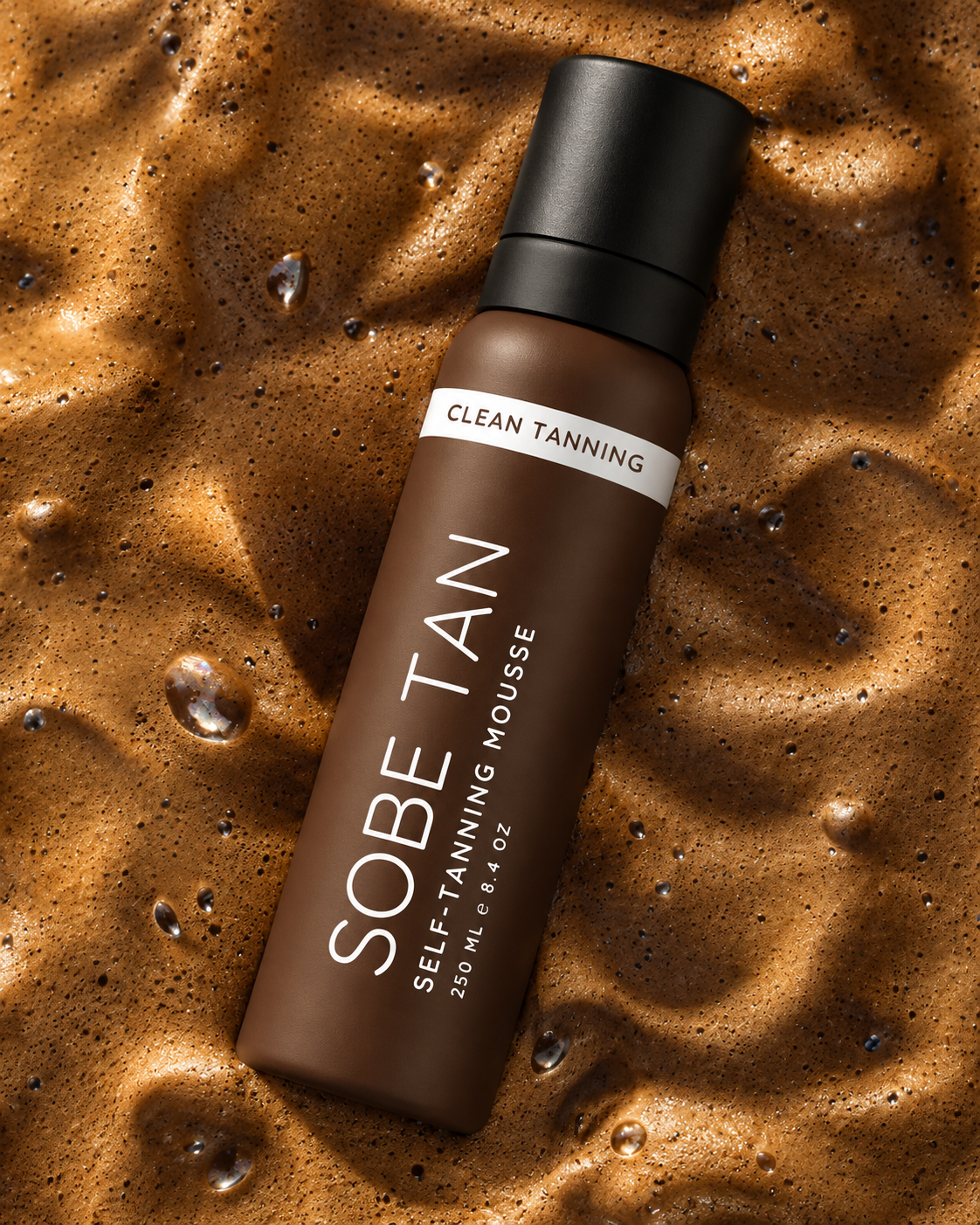 SoBe Tan self-tanning mousse bottle lying diagonally on textured brown foam.